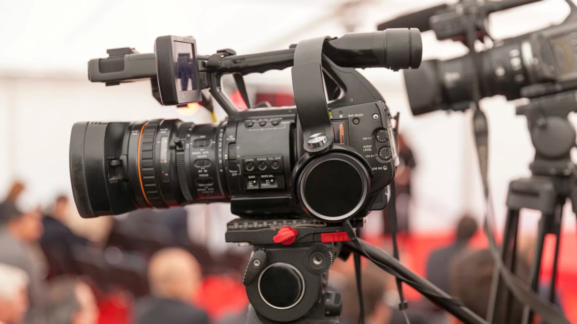 Professionelle Videokamera auf einem Stativ gerichtet auf eine Medien-/Presseveranstaltung mit unscharfem Hintergrund.